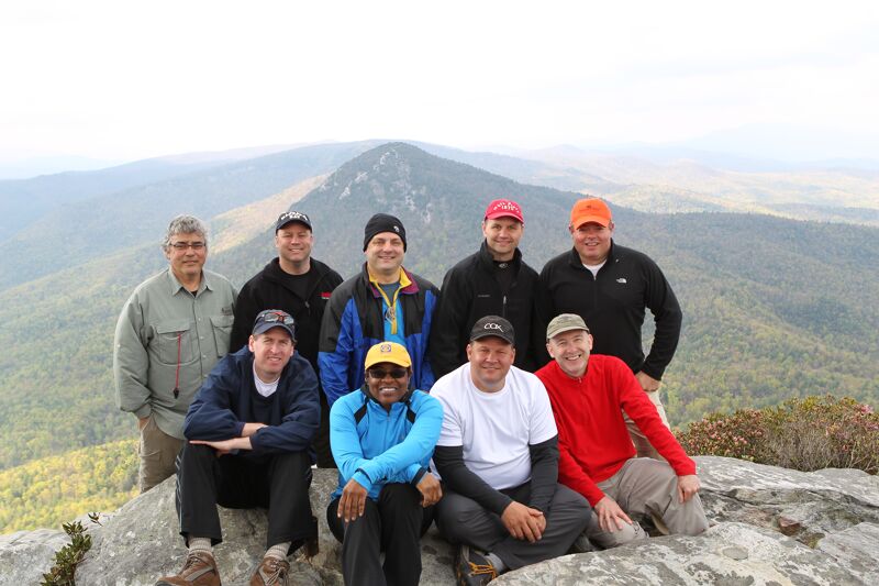 A group of nine men are posing for a photo on a rocky mountaintop. They are dressed in casual outdoor clothing, including jackets and hats. The background features a scenic view of rolling hills and mountains under a bright sky. The overall atmosphere suggests a hiking or outdoor adventure.